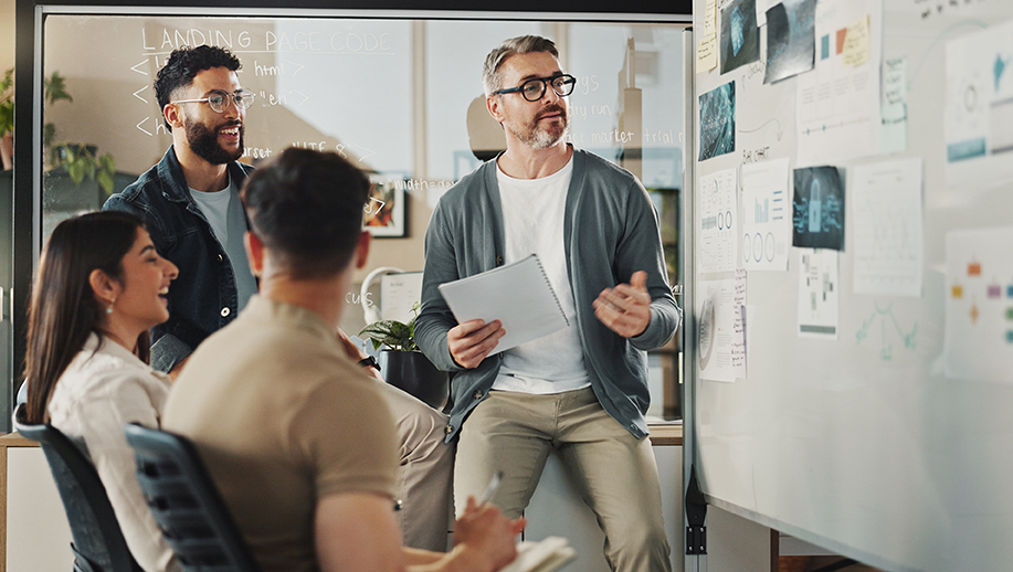 Two woman in an office standing in front of a glass whiteboard discussing sticky notes that are on the board.