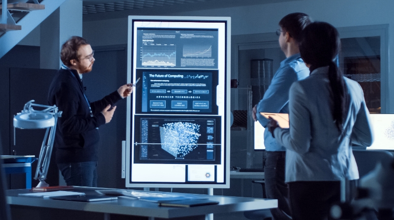 A woman working next to two men all sitting at desks with computers, analyzing data, testing approaches and researching solutions.